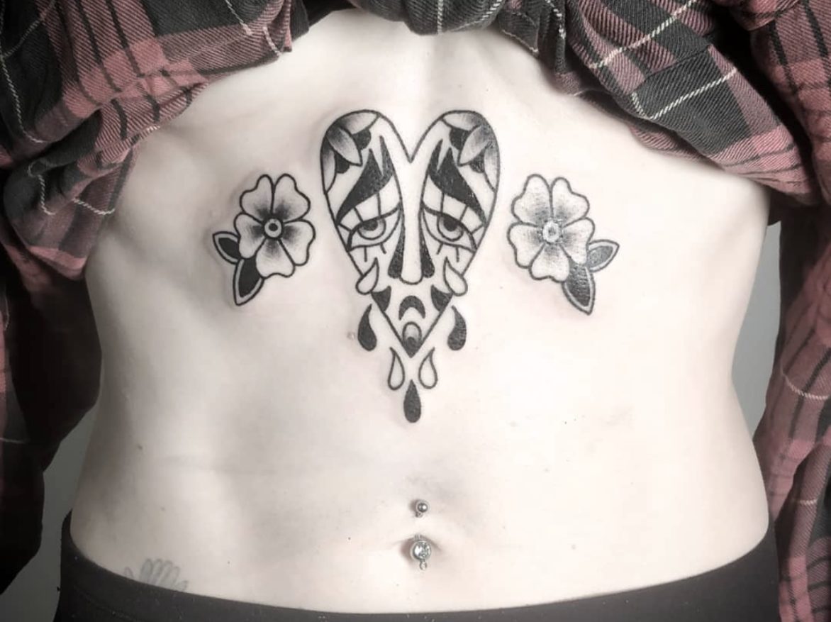 traditional blackwork flash design of heart with face inside crying tears and two black and grey flowers on either side tattooed on stomach above belly button