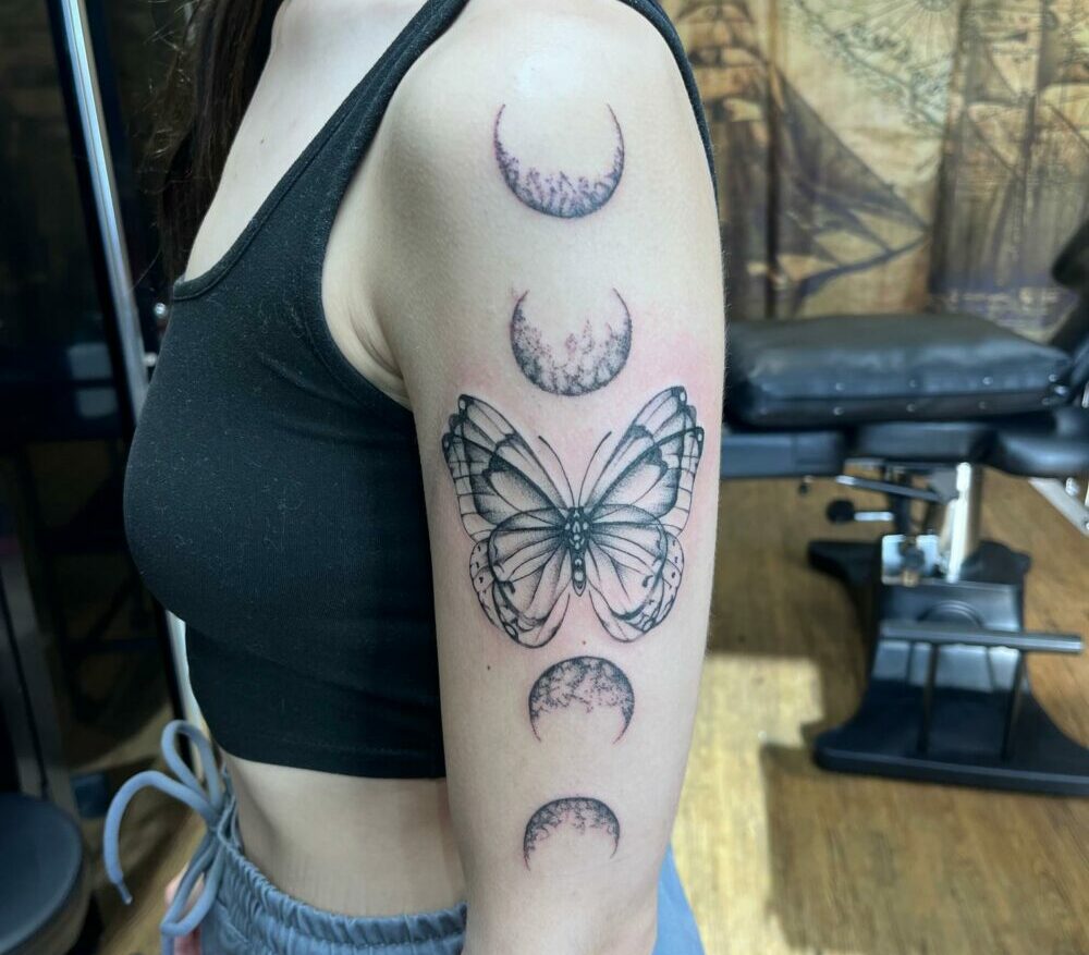 dotwork butterfly tattoo with phases of the moon above and below it, tattooed on outside left arm