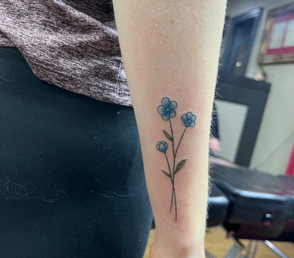 Fineline tattoo of three blue flowers with stems tattooed on the outside of a wrist